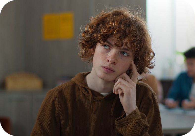 Un ragazzo adolescente con capelli ricci rossi, seduto in classe, guarda in alto pensieroso con la mano sul viso.