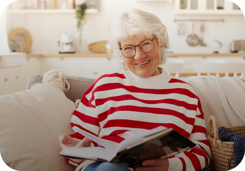 Una donna anziana con occhiali e maglione a righe rosse e bianche è seduta sul divano in salotto e sorride mentre legge un libro.