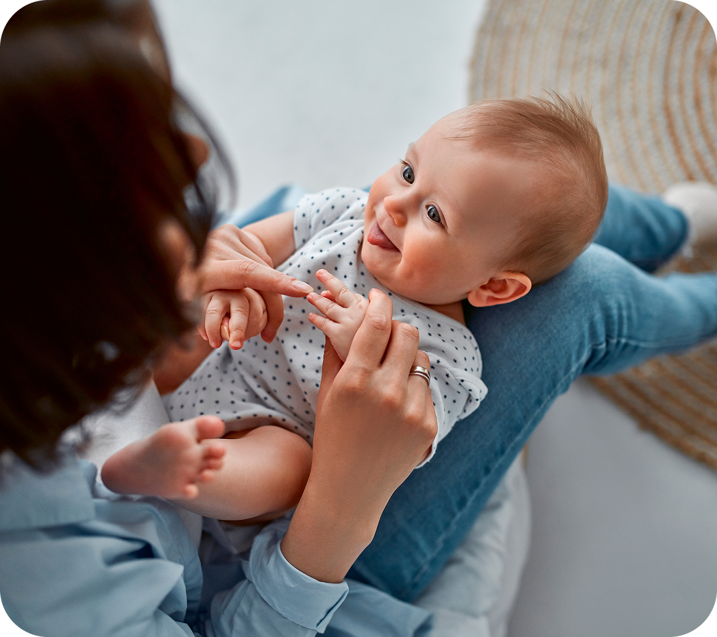 Un neonato in body bianco sorride guardando la madre che lo tiene in braccio e gli accarezza le mani, seduta su un tappeto.